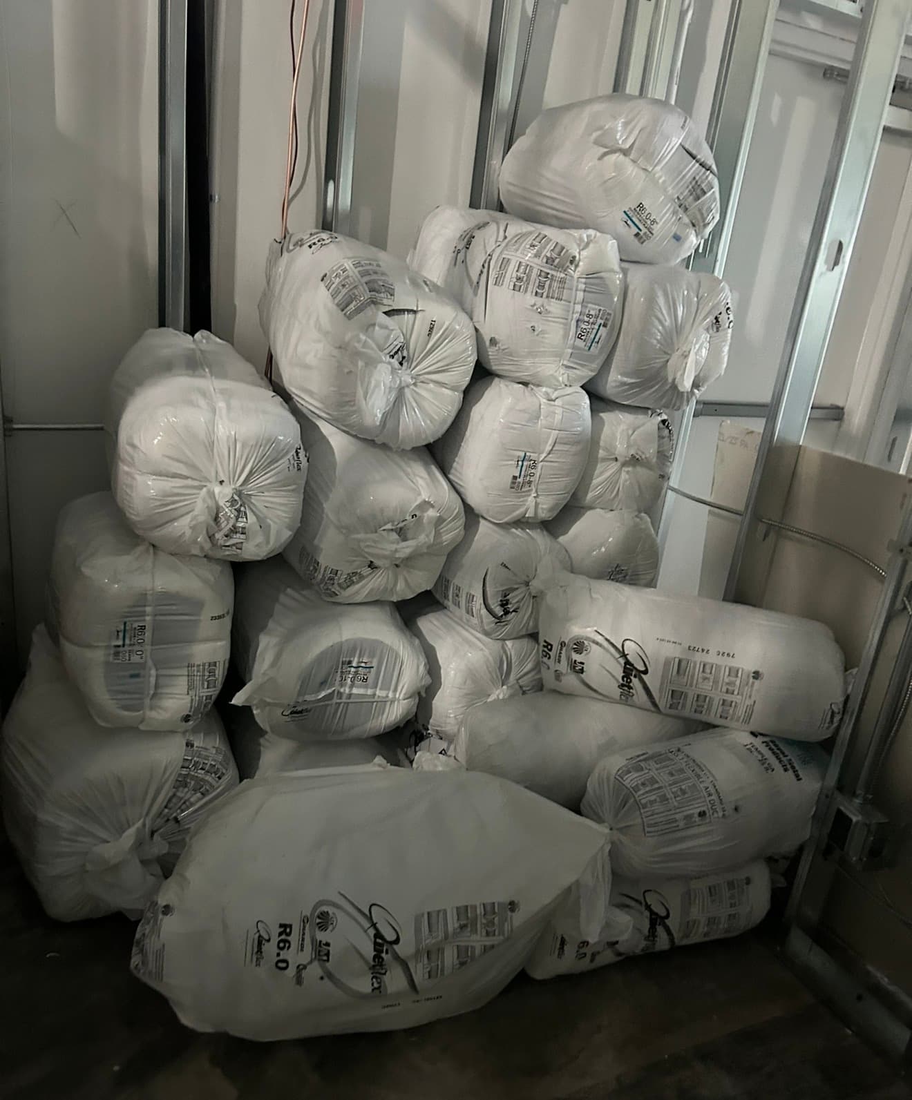 Stacked rolls of white plastic-wrapped flexible air duct insulation against metal wall studs.